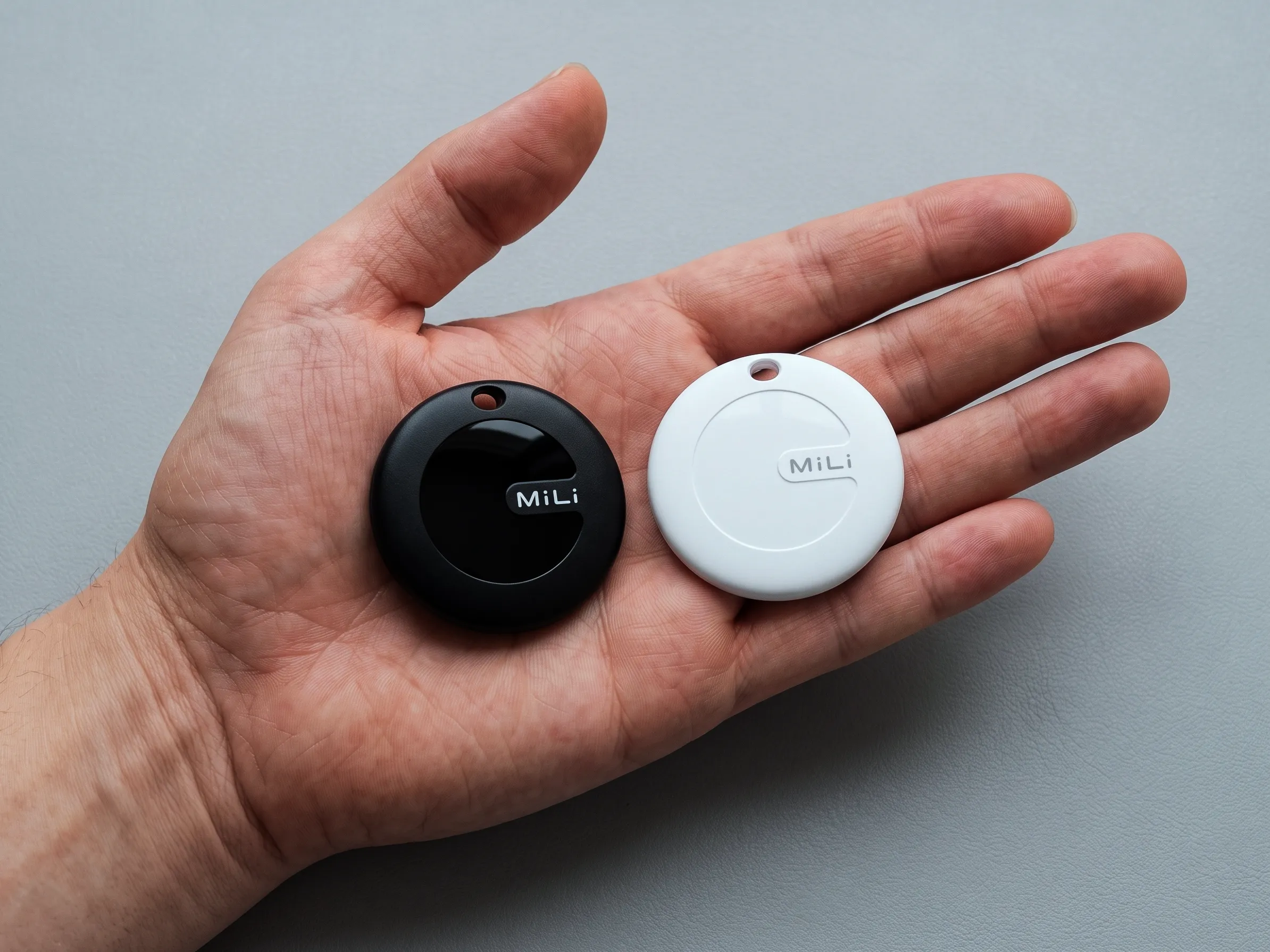 Los dos trackers MiLi MiTag Duo, el negro y el blanco, apoyados en la palma de una mano adulta: cada disco mide algo menos de la mitad del ancho de la palma