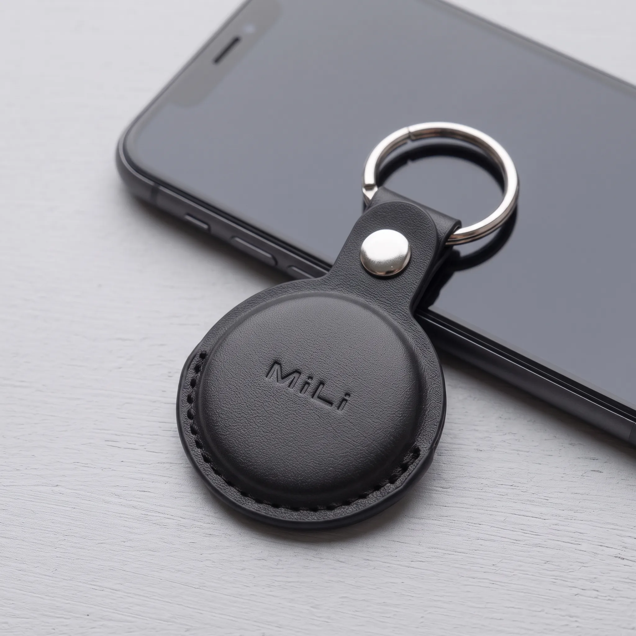 Tracker MiLi MiTag Duo en su funda de cuero negra, apoyado contra el borde de un teléfono con la pantalla apagada